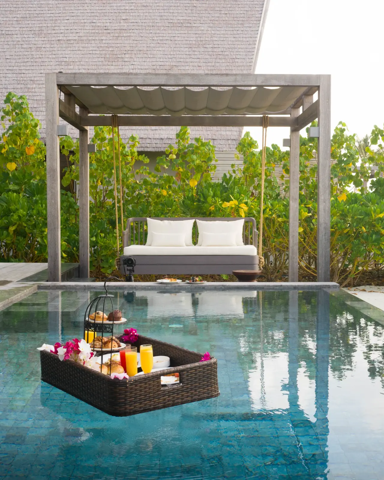 A floating breakfast in a wicker tray on a private pool, with the swing and the greenery behind.