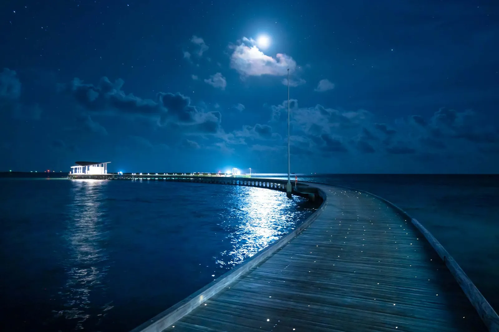 The water villas’ jetty at night, lit up, under the moon and the clouds.