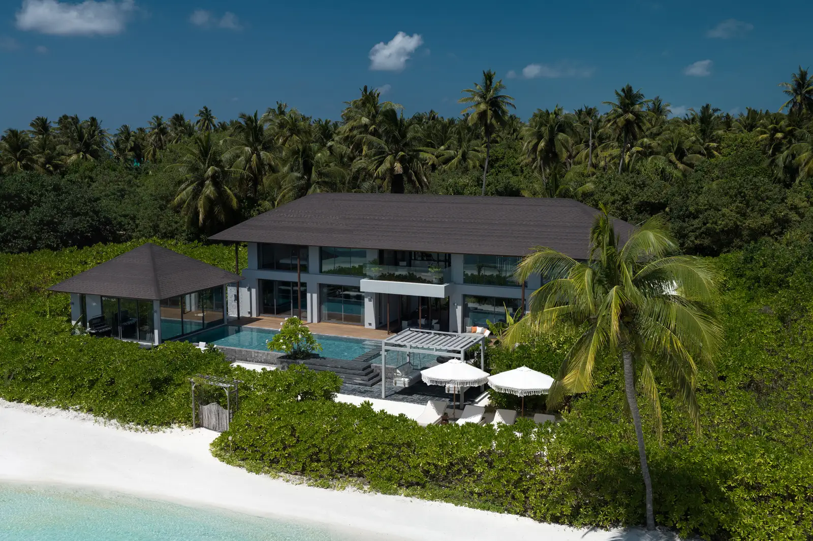 Bella Six from above: the 2-storey villa among the palms, its pools, the pavilion and the parasols on the beach.