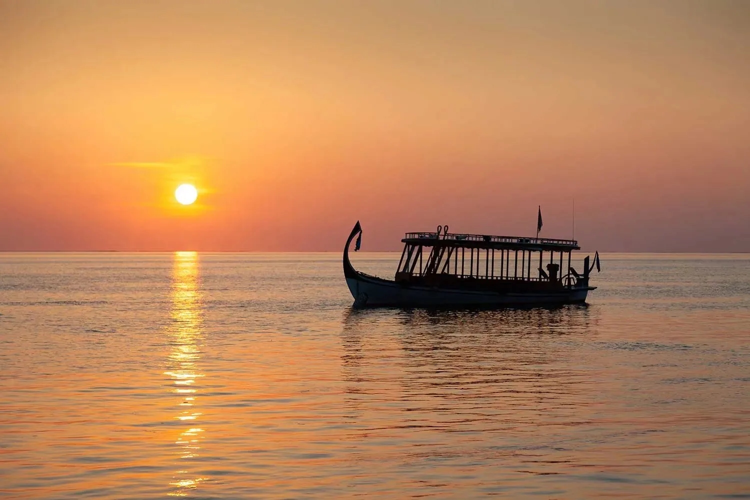 A Maldivian dhoni on the sea at sunset, the sun low on the horizon.