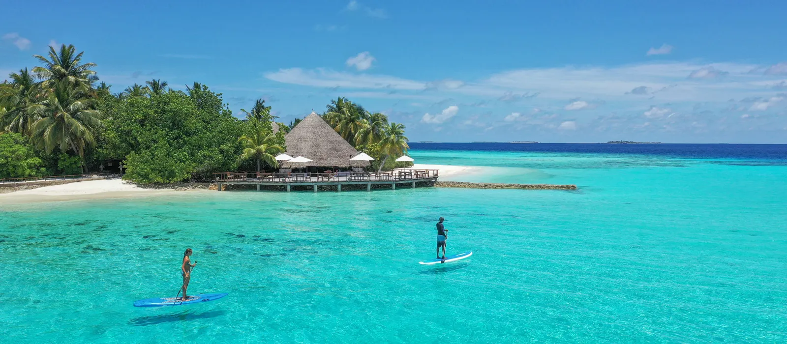 2 paddleboarders on the turquoise lagoon, the bar on its jetty and the island’s trees behind.