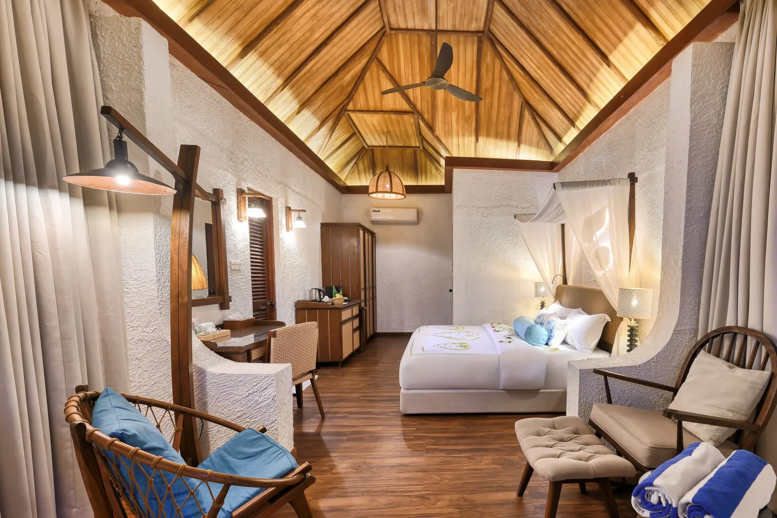 A Deluxe Beach Bungalow bedroom: the bed under the pitched timber roof, a rattan armchair and the desk.