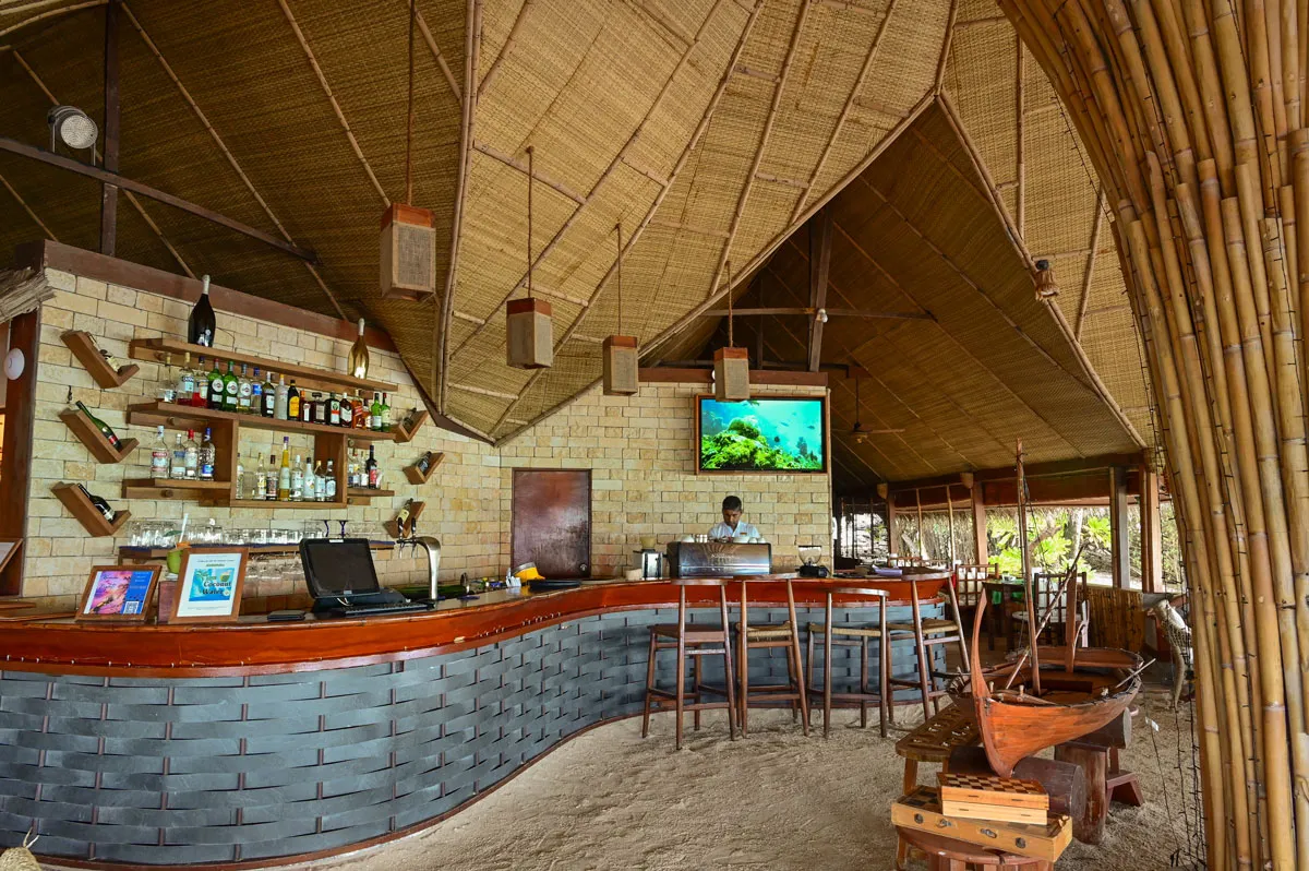 The Sand Bar’s counter on the sand under its thatched roof, with stools and bottles on the shelves.