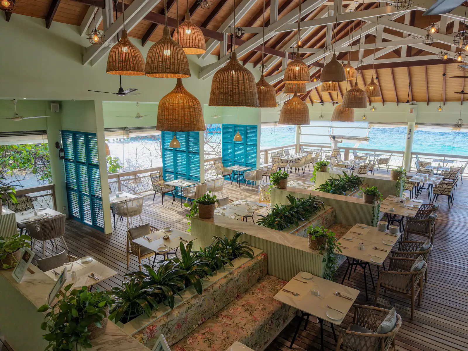 Maaga’s dining room from above: wicker lamps under the timber roof, blue shutters and the lagoon beyond the tables.