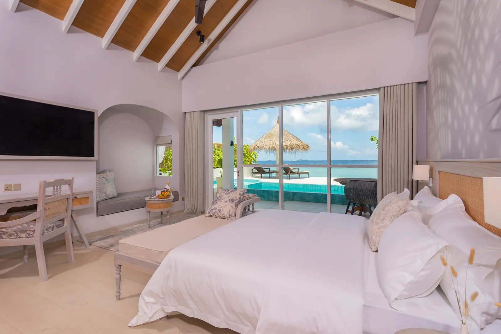 A Sunrise Beach Pool Villa: the bed under the pitched roof and, beyond the glass, the pool, a thatched parasol and the sea.