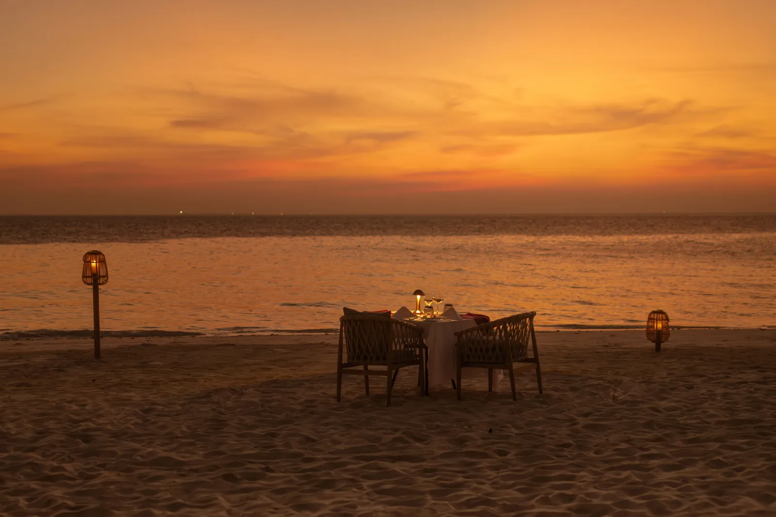 A table for 2 on the sand at sunset, between 2 lanterns, under an orange sky over the sea.