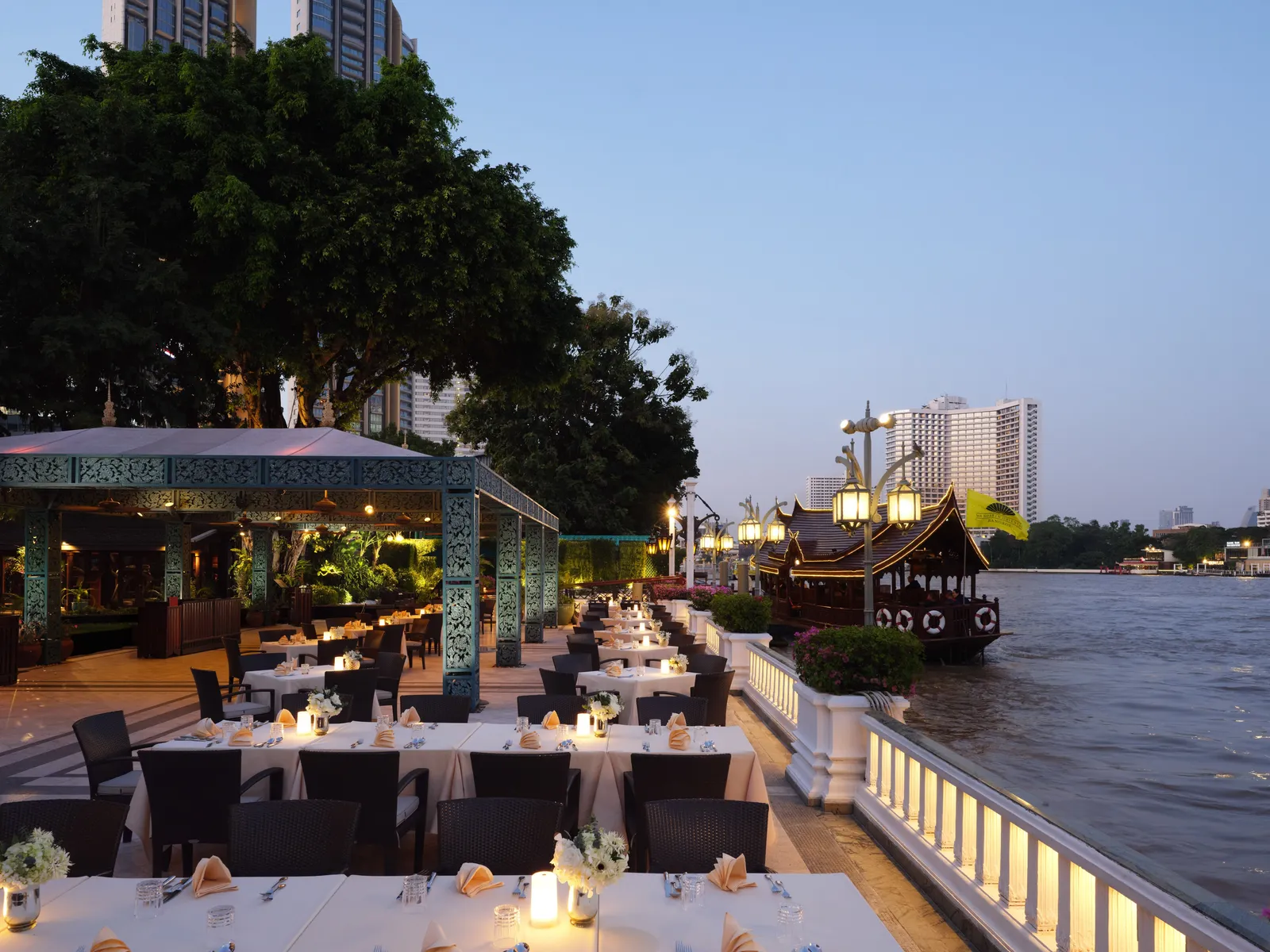 Tavoli apparecchiati sul Chao Phraya all’imbrunire, con una barca di teak dell’hotel ormeggiata accanto