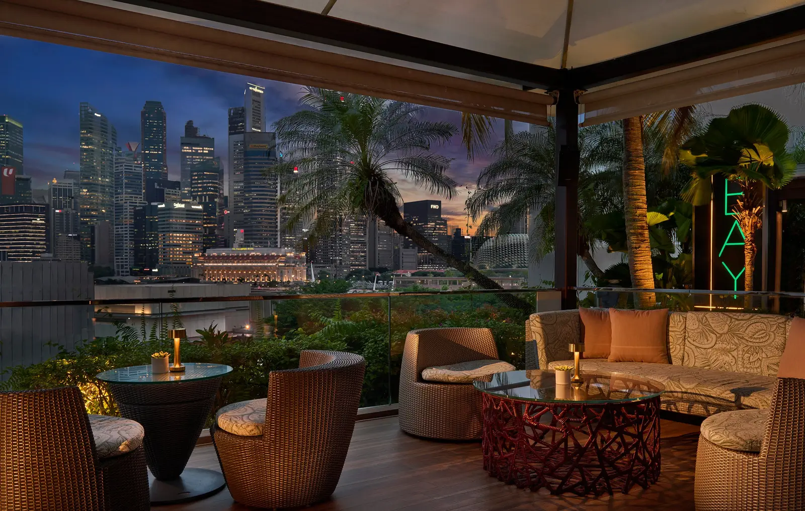 BAY@5 at dusk: wicker sofas and armchairs under the canopy, with the lit skyline beyond the palms.