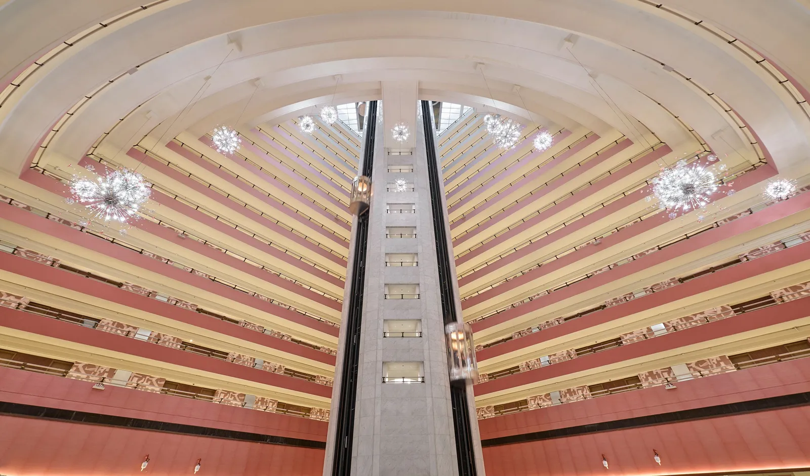 The atrium seen from below: tiers of pink and cream balconies fanning out, 2 glass lifts at the centre and globe chandeliers hanging between them.