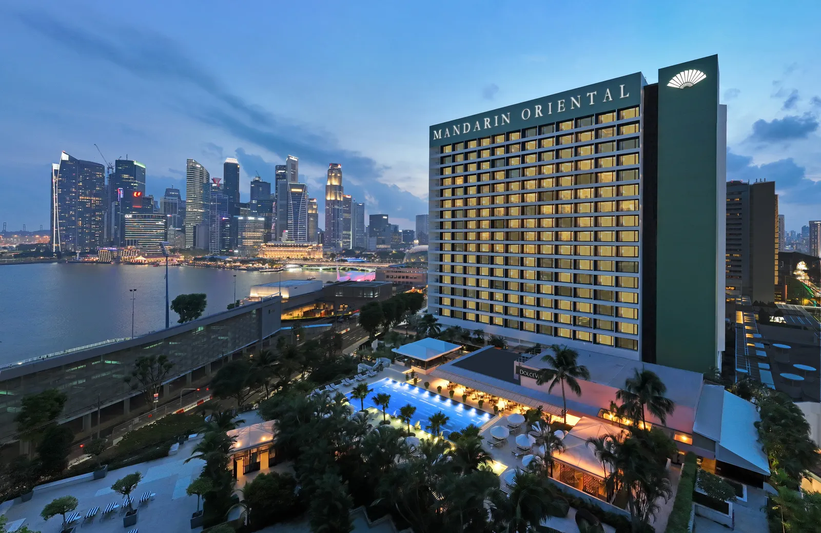 The hotel at dusk: the façade with its rooms lit and the Mandarin Oriental sign, the pool among palms at its foot and, on the left, the towers across the bay.