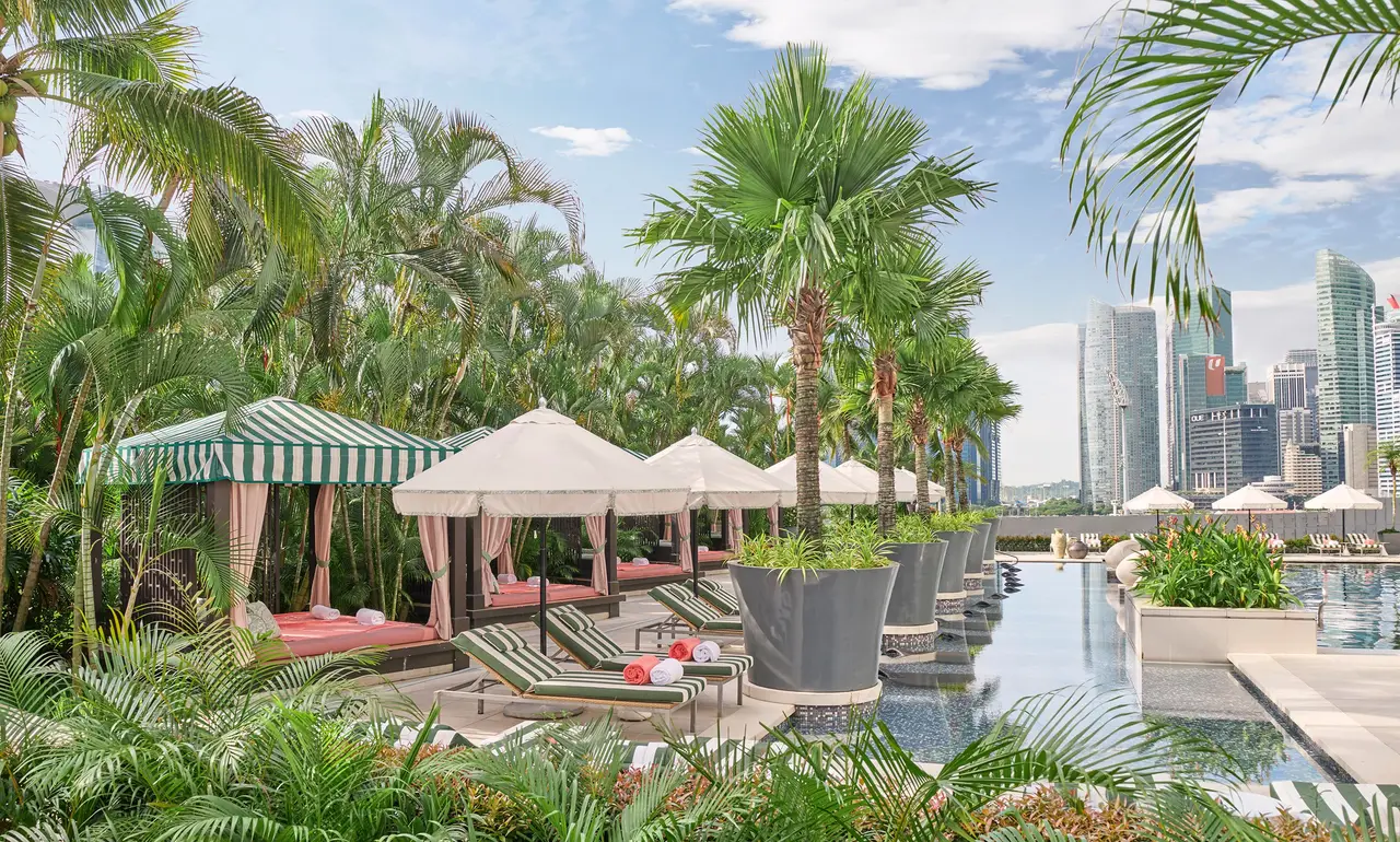The pool by day: cabanas with green-striped roofs, white parasols, loungers and palms, with the financial district’s towers behind.