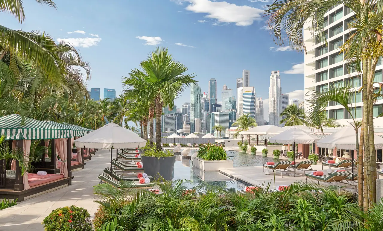 The pool among the palms: striped loungers, parasols and cabanas along the water, and the towers beyond its edge.