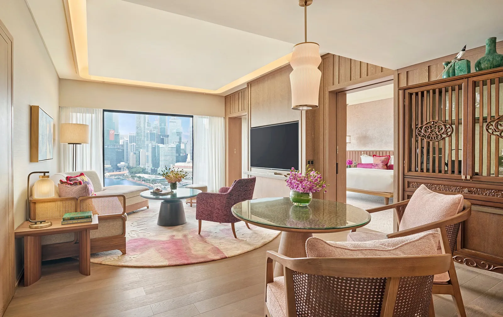 The living room of the Two-Bedroom Premier Marina Bay Suite: a round table, a sofa and the bedroom with fuchsia cushions through the door, with the bay in the window.