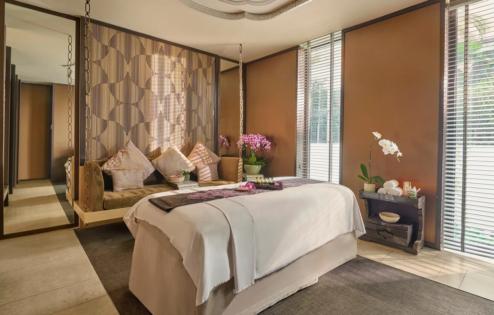 A spa treatment room: the massage bed, a daybed hung on chains with cushions and orchids, light coming through the blinds.
