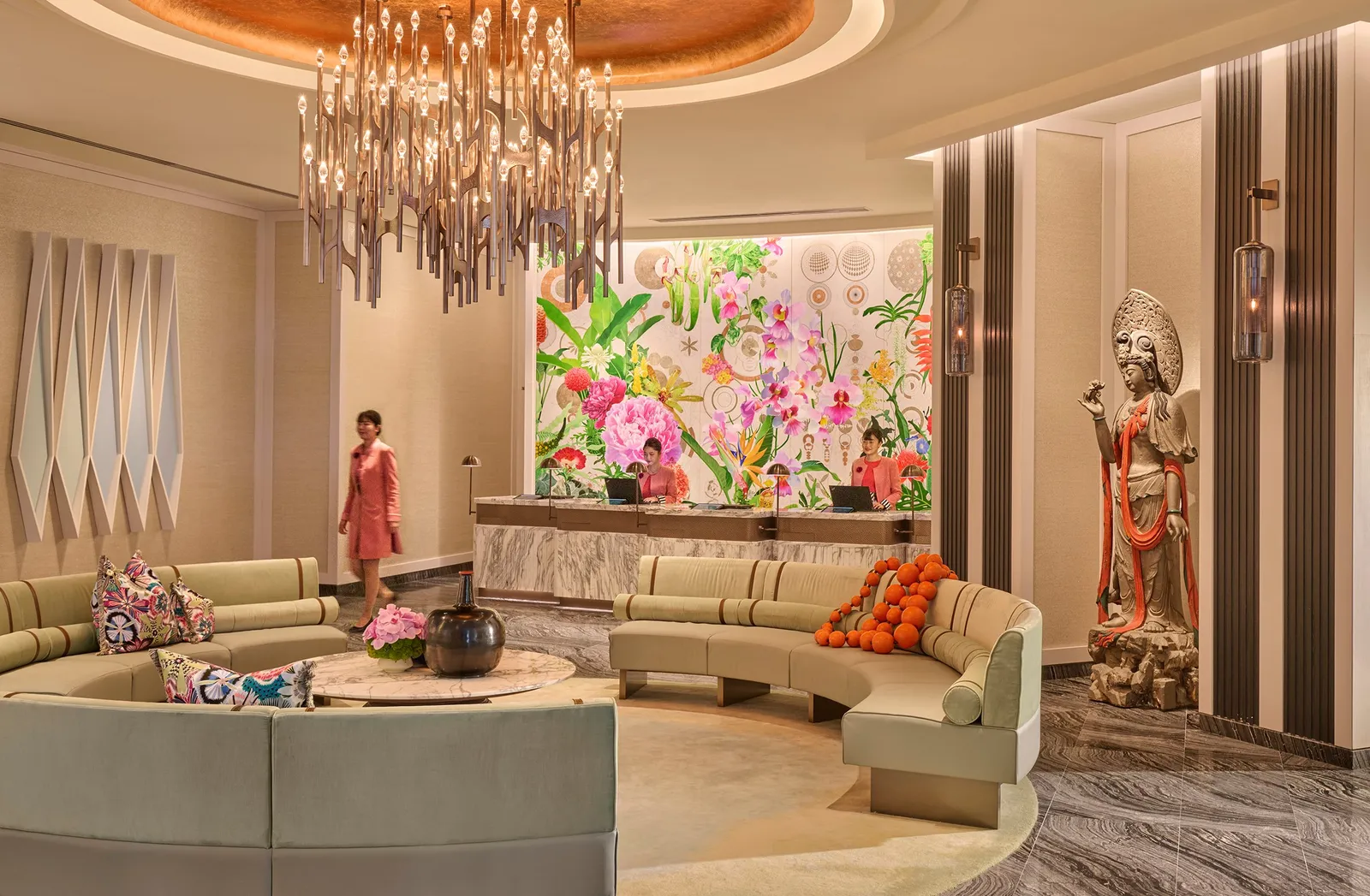 The front desk under a mural of tropical flowers, with a cascading chandelier, curved sage-green sofas and a painted wooden statue.