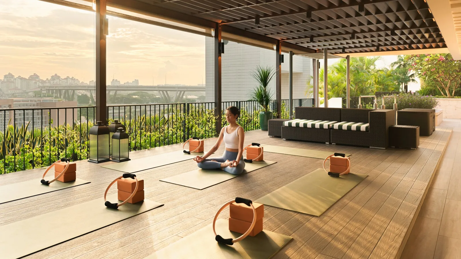 The Wellness Terrace: a woman meditating on a mat, yoga props on the wooden floor and the city in morning light.