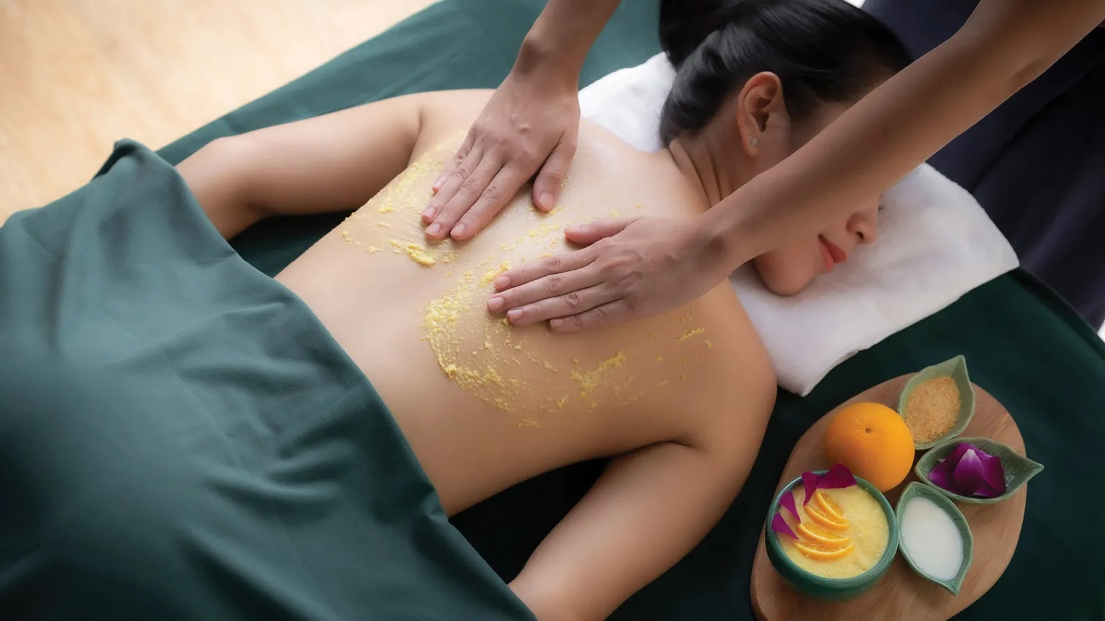A massage at Banyan Tree Spa: 2 hands on the back of a guest lying down, a tray of fruit and scrubs beside her.
