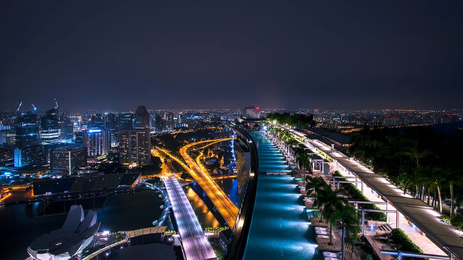 The SkyPark at night from above: the pool lit blue between the palms, the roads and towers alight below.