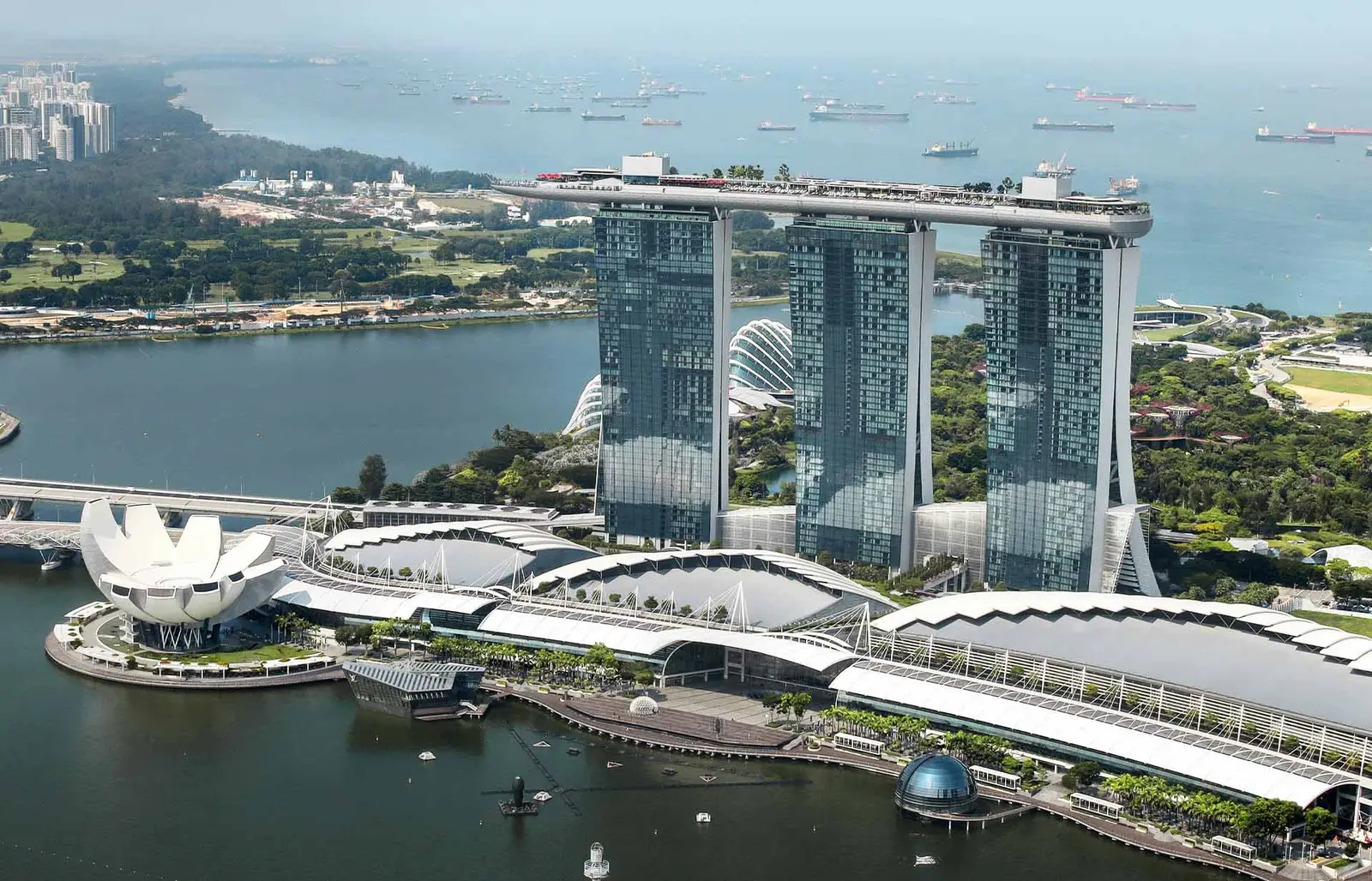 The 3 towers of Marina Bay Sands with the SkyPark on top, from the air: the white lotus-shaped museum on the bay in front, the gardens and ships at anchor in the strait behind.