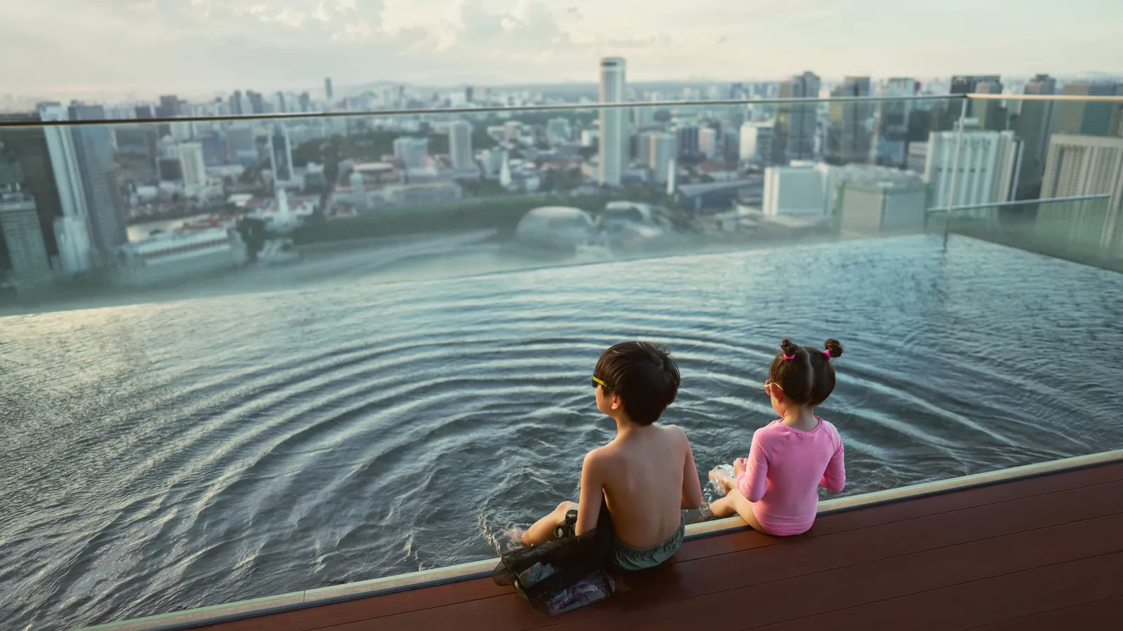 2 children sitting on the pool’s edge look at the city beyond the glass parapet.