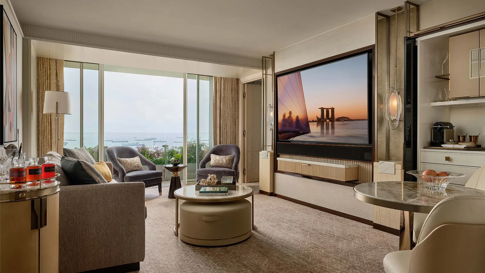 Sands Bay Suite: the living room with a sofa, a round table and a wall-mounted television, the window on the sea at the far end.