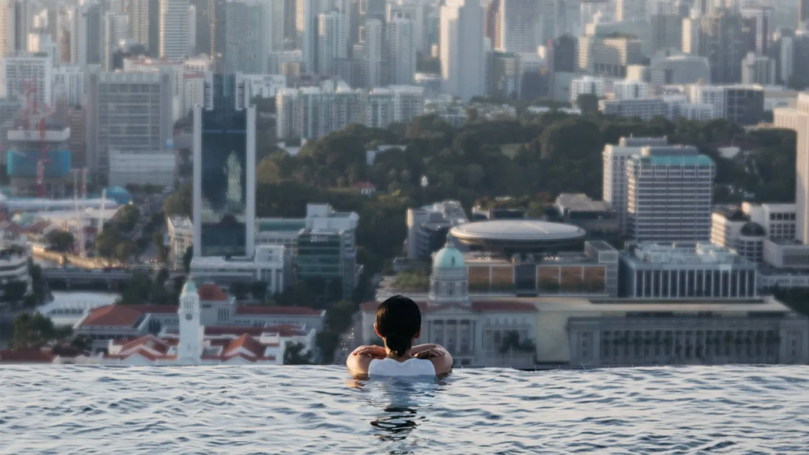 A guest at the pool’s edge looks out at red roofs, a clock tower and a dome in the old centre, under the office towers.