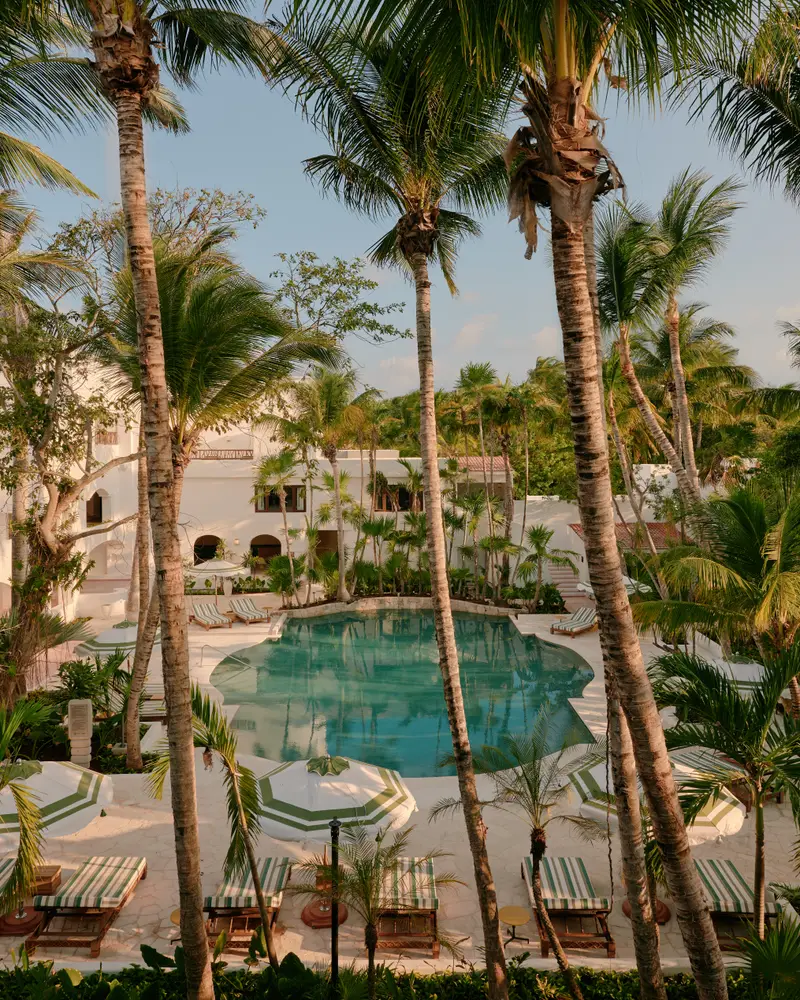 La piscina verde fra le palme, con lettini, ombrelloni e gli edifici bianchi sullo sfondo