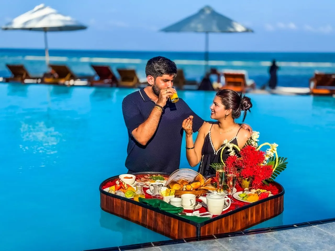 A couple in the pool with a heart-shaped floating breakfast tray, and loungers under parasols behind.