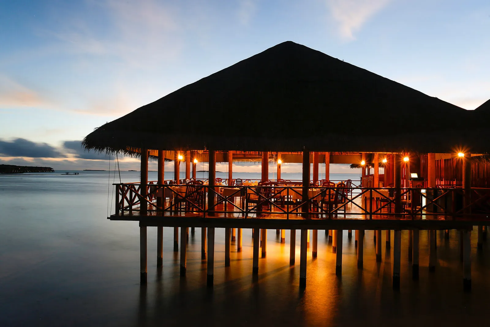 The over-water pavilion at dusk, lit beneath its great thatched roof, its stilts reflected in the lagoon.