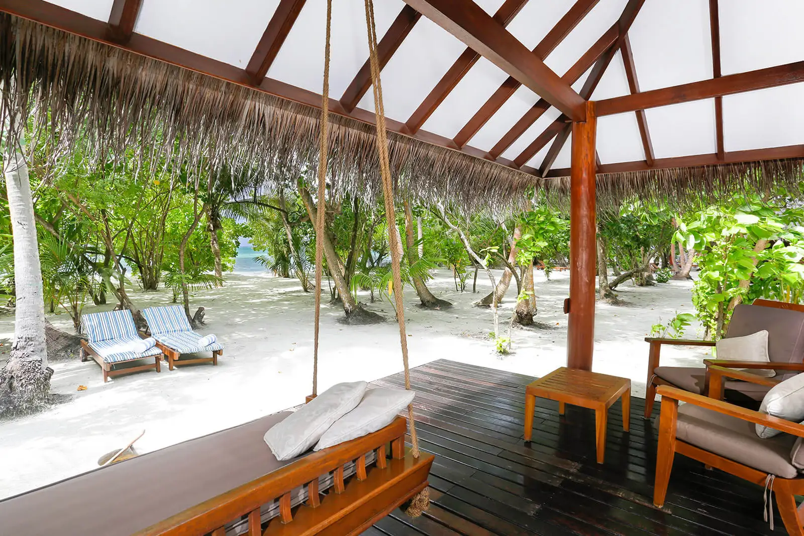 A beach villa’s covered deck, with its swing seat and armchairs, and the sand among the trees in front.