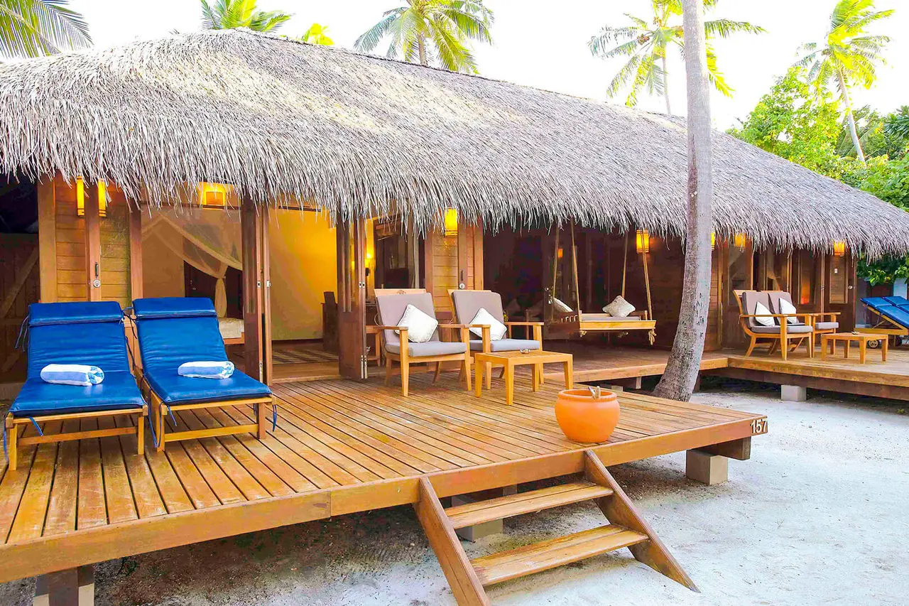 A beach suite’s wooden deck under the thatched roof, with 2 blue loungers and seating in front of the rooms.