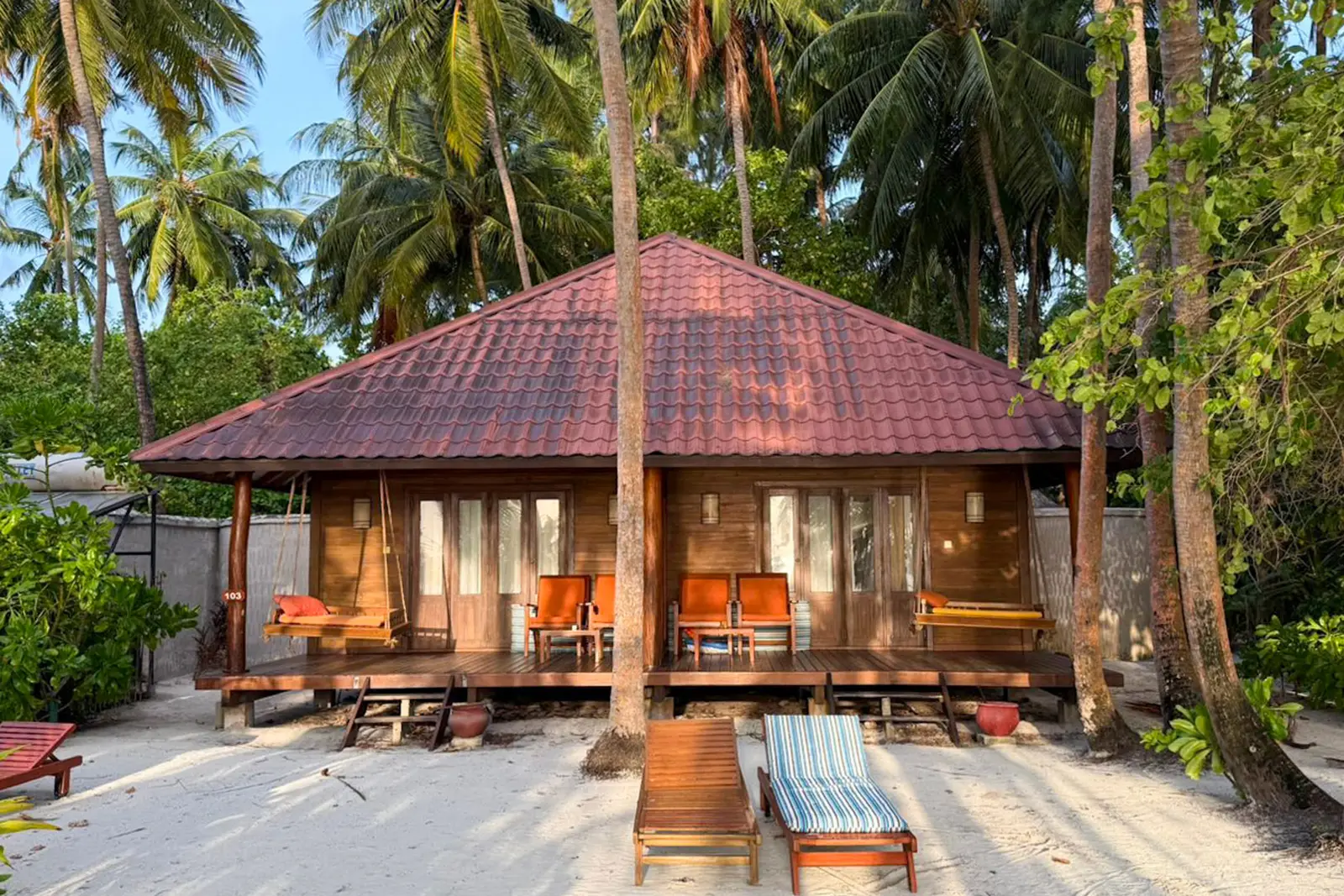 A semi-detached beach villa in timber with a red roof, its veranda and loungers on the sand among the palms.