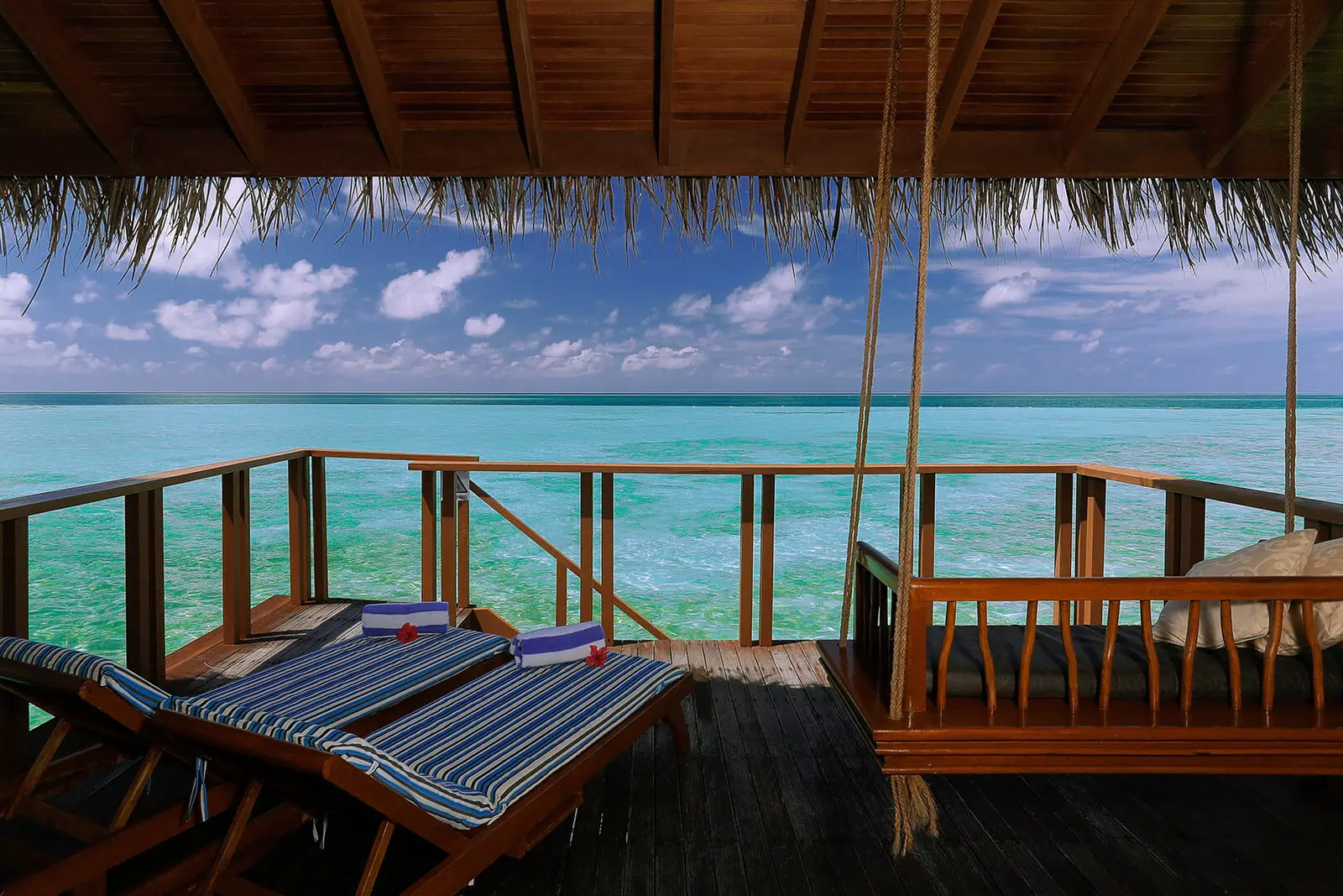 A water villa’s deck, with striped loungers, the swing seat and the turquoise lagoon beyond the railing.