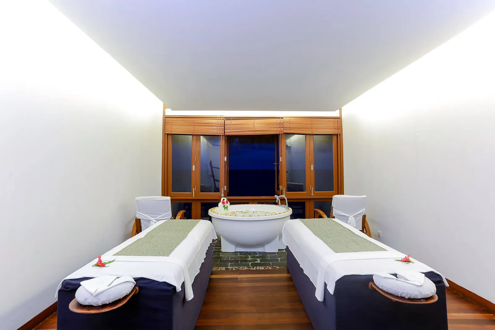 A spa suite with 2 massage beds, a round bath of flowers between them and the glass doors open to the sea.