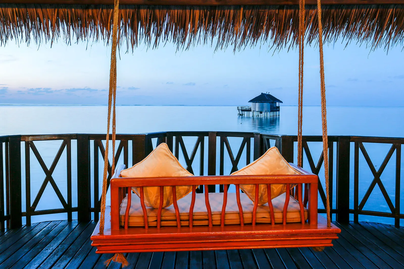 The cushioned wooden swing on a water suite’s deck at dawn, with a distant pavilion in the lagoon.