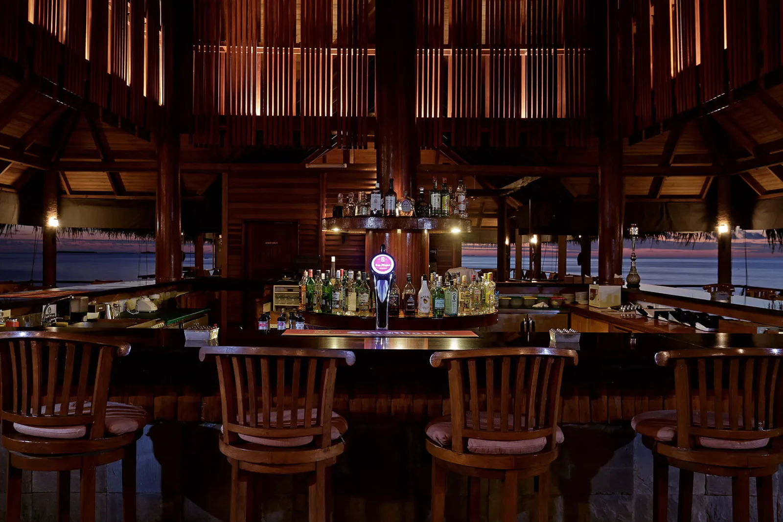 The Vilu Bar counter with its wooden stools, the lit bottles and the pavilion’s slatted walls.