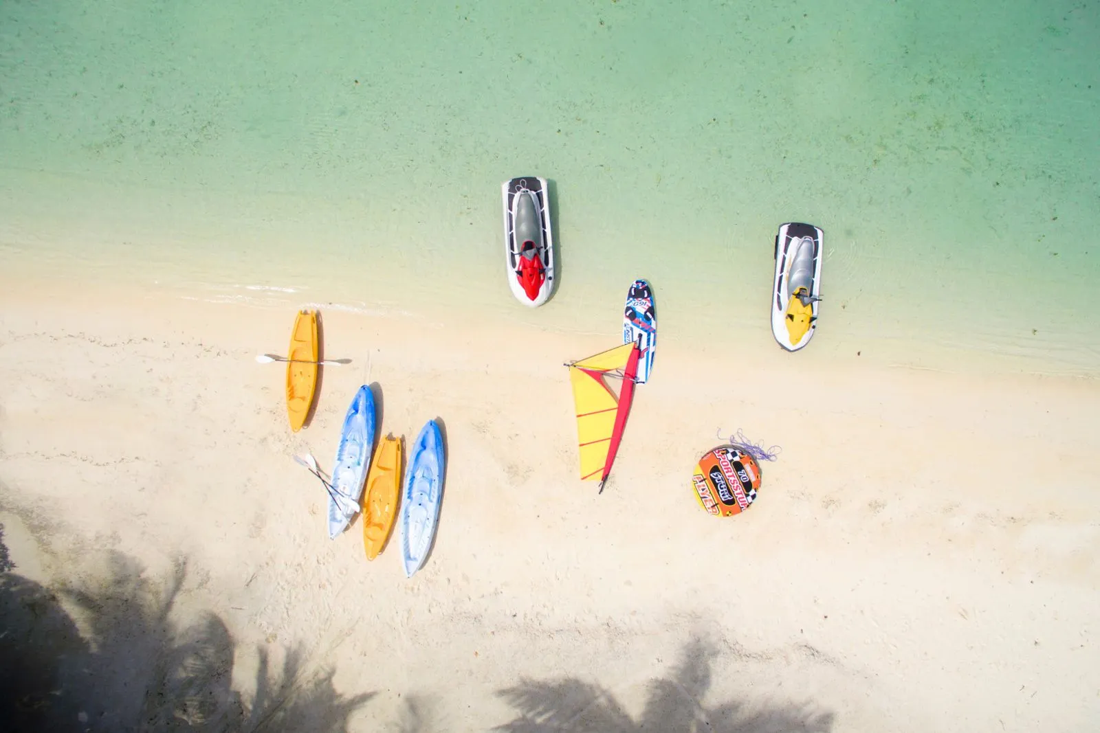Jet skis, kayaks, boards and an inflatable ring on the white sand, seen from above beside the shallow water.