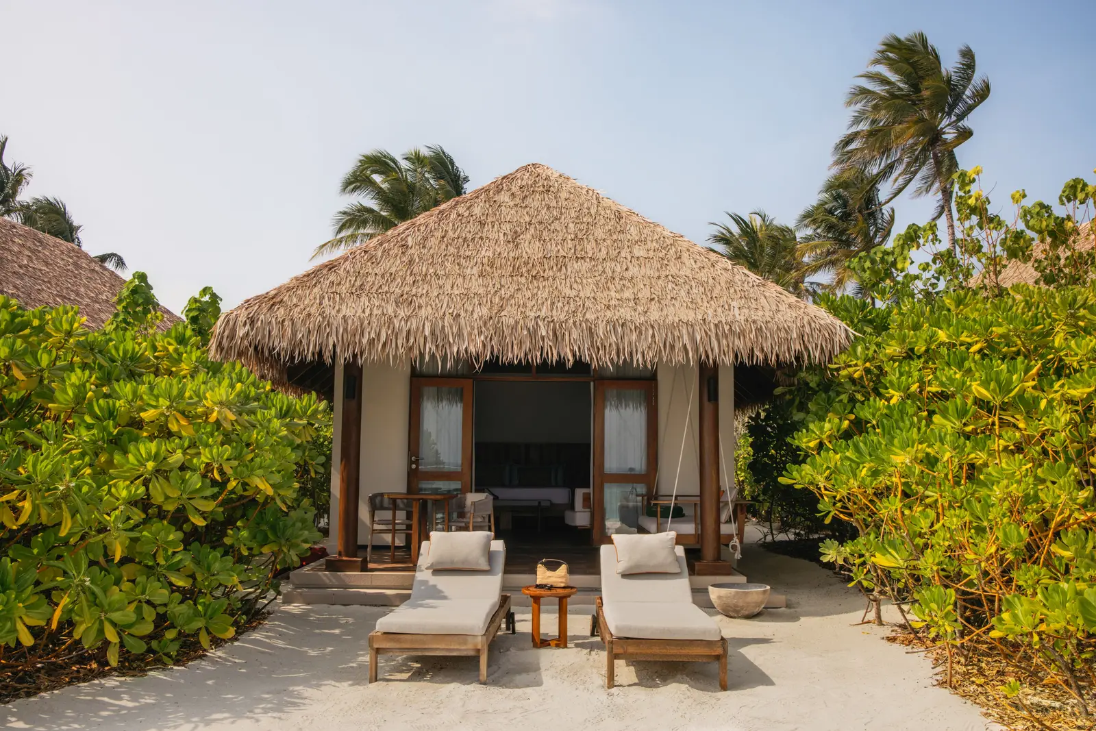 A Sunset Beach Villa under its thatched roof among the greenery, 2 loungers and a parasol on the sand in front.