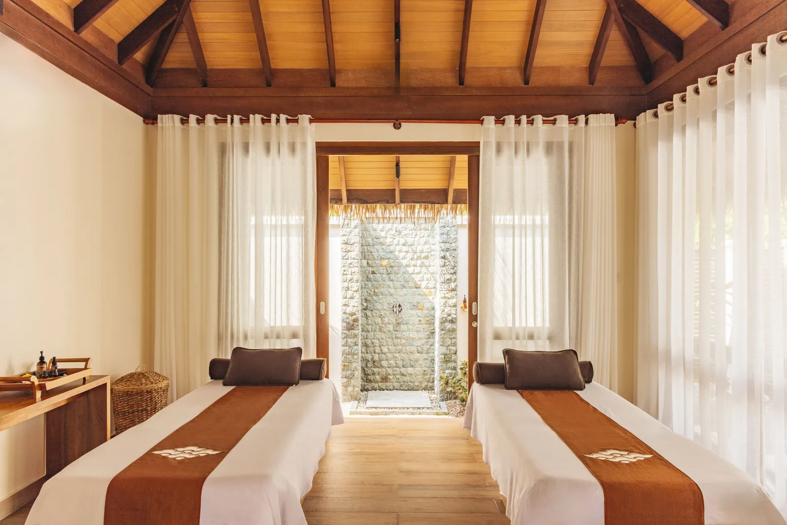 A couples’ treatment room at the spa: 2 beds with orange throws and the door open onto the outdoor shower.