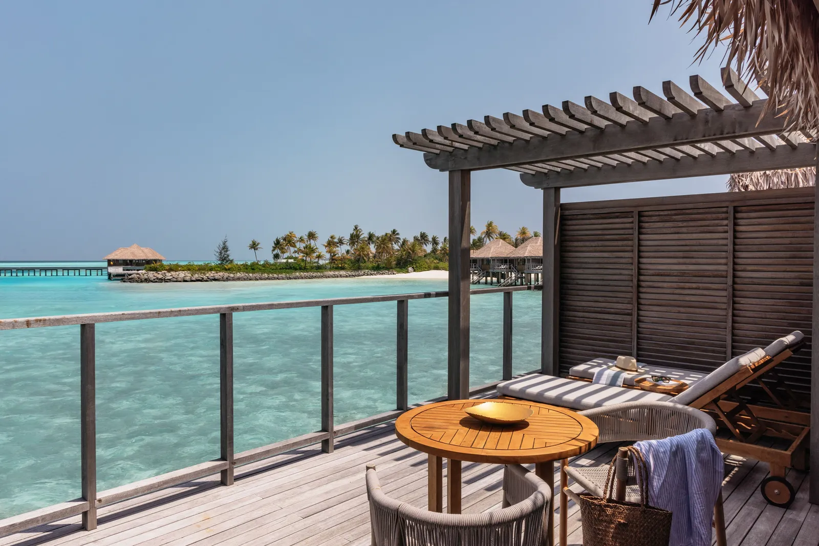 A water villa’s deck under its pergola, with the round table, the loungers and the sea to the horizon.