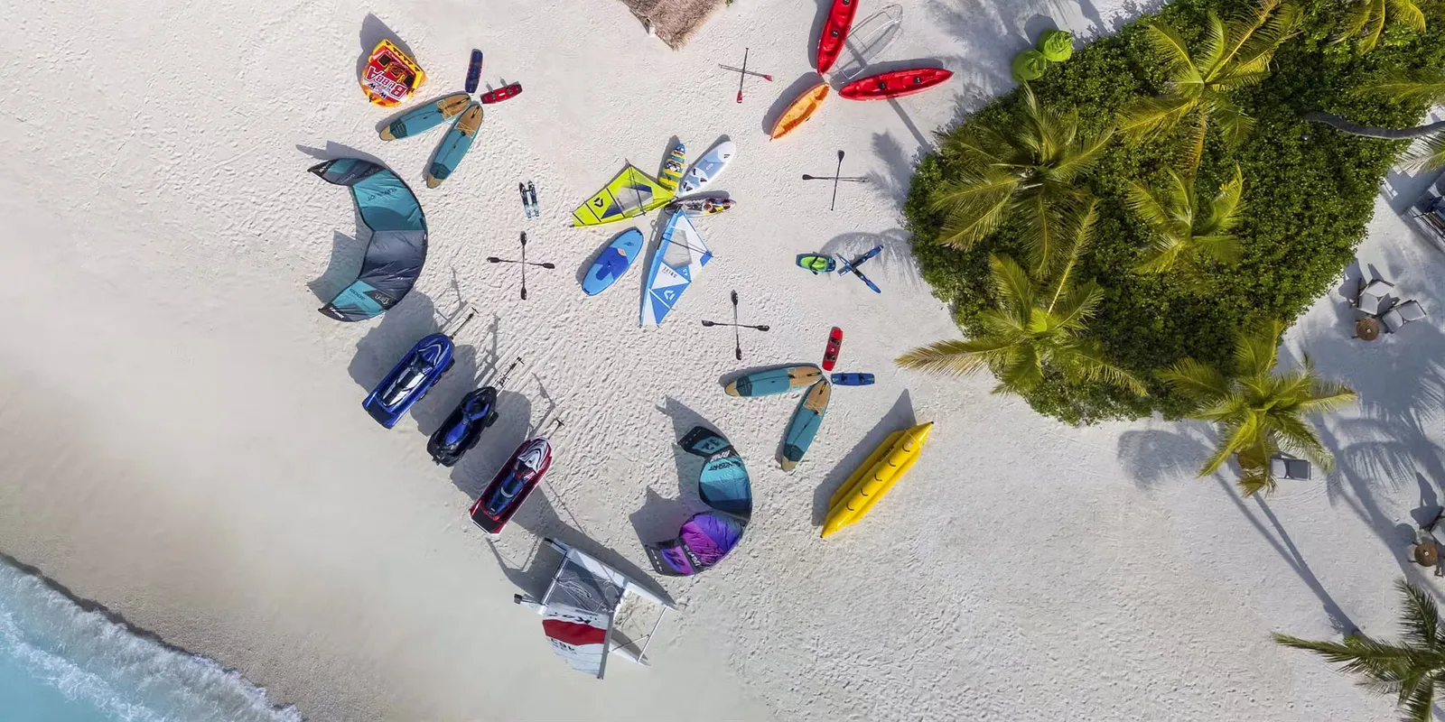 From above, on the sand: kayaks, paddleboards, sails and a jet ski at the water-sports centre, beside the greenery.