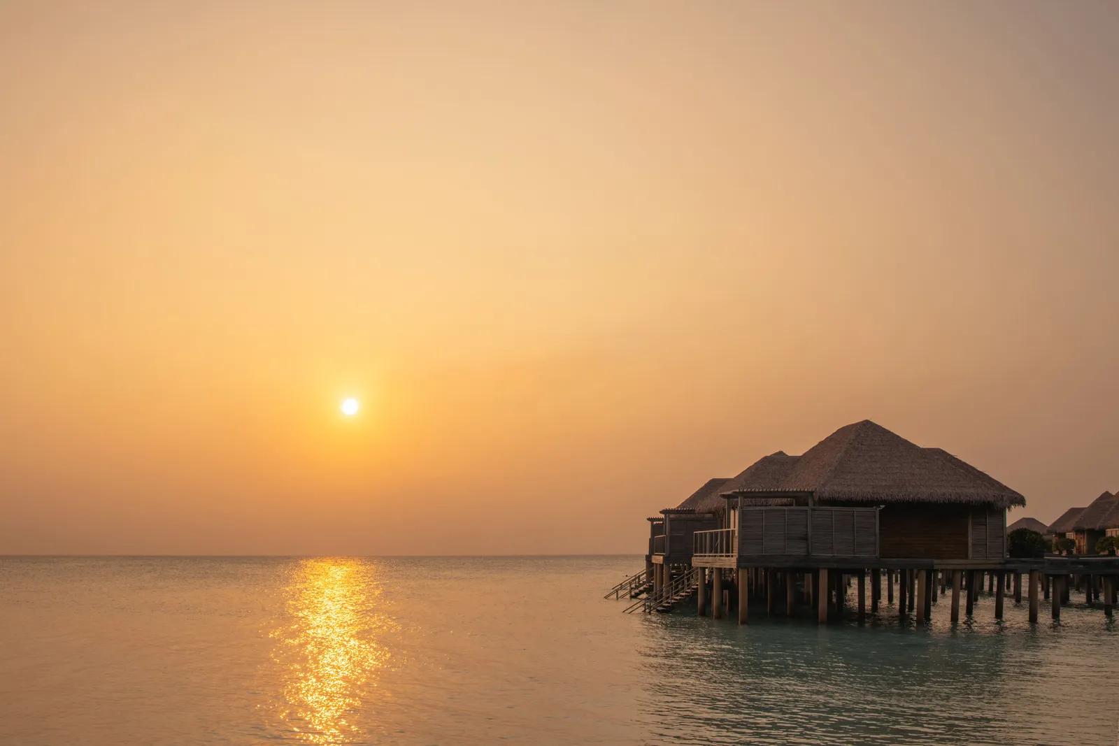 The sun setting over the sea beside 2 thatched water villas.