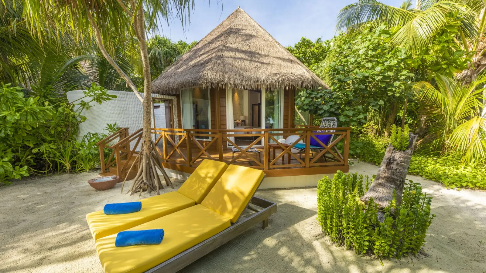 A thatched Beach Villa among the palms, its veranda and 2 yellow loungers on the sand.