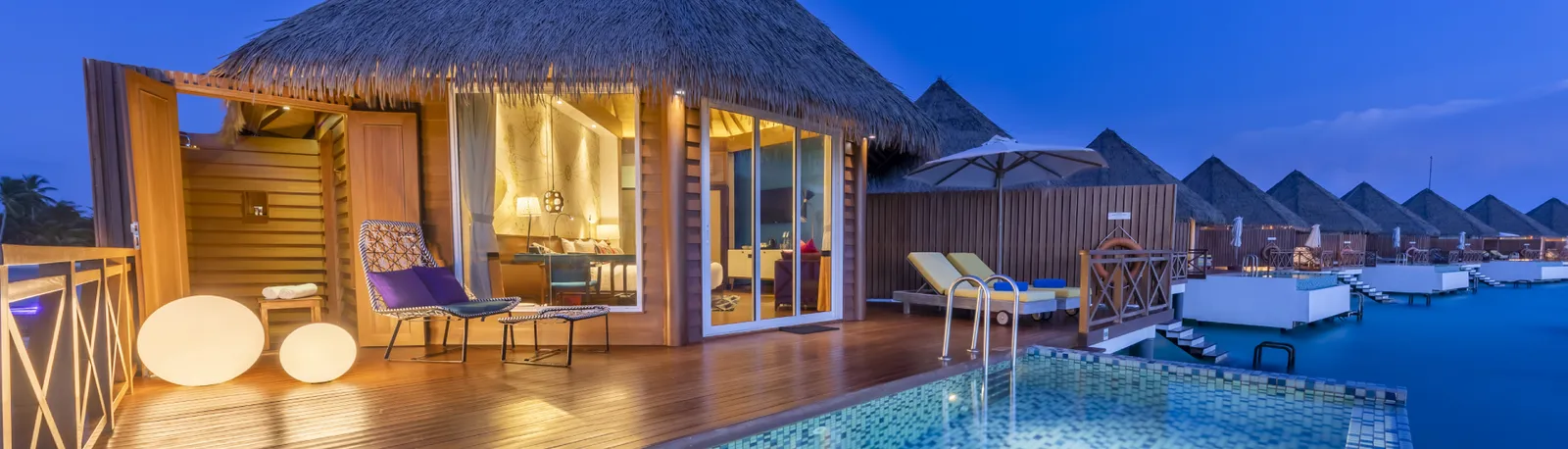 The deck of an Overwater Sunset Pool Villa in the evening, with its pool, loungers and lights on.