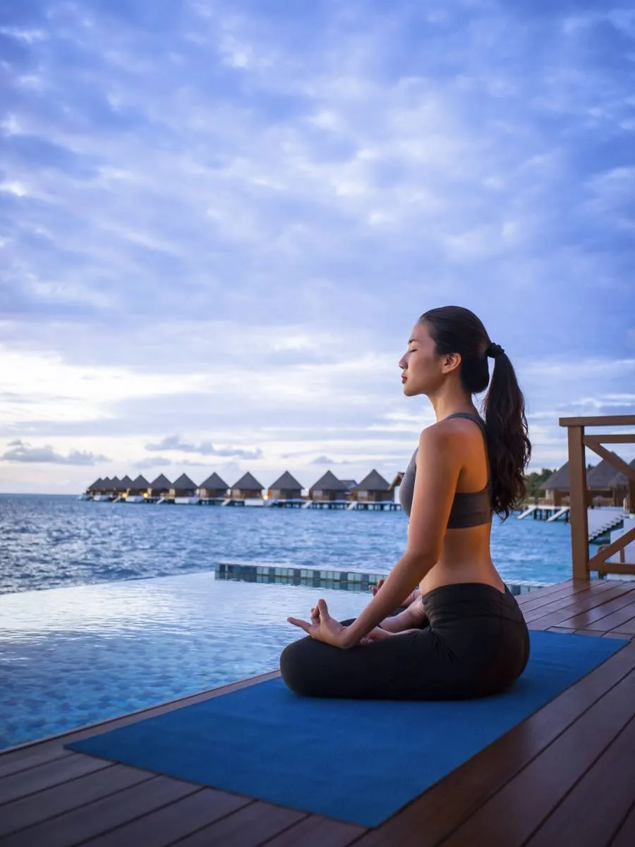 A woman meditating on a mat on the deck of a water villa, beside its pool.