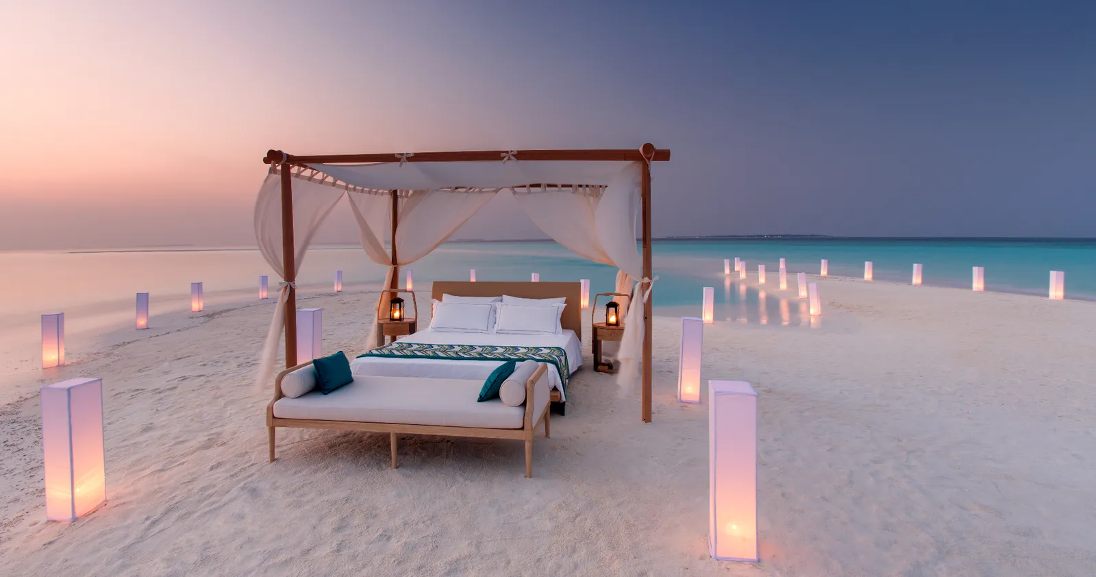 A four-poster bed on the sandbank, lanterns lit at sunset