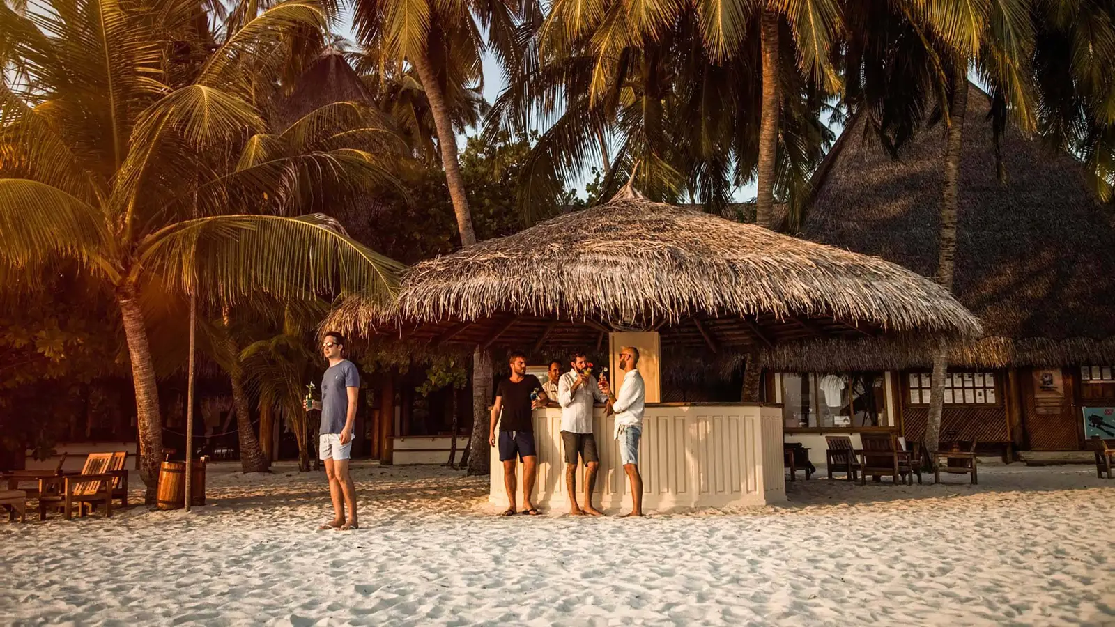 The Beach Bar under its thatched roof on the sand, a few guests standing among the palms.