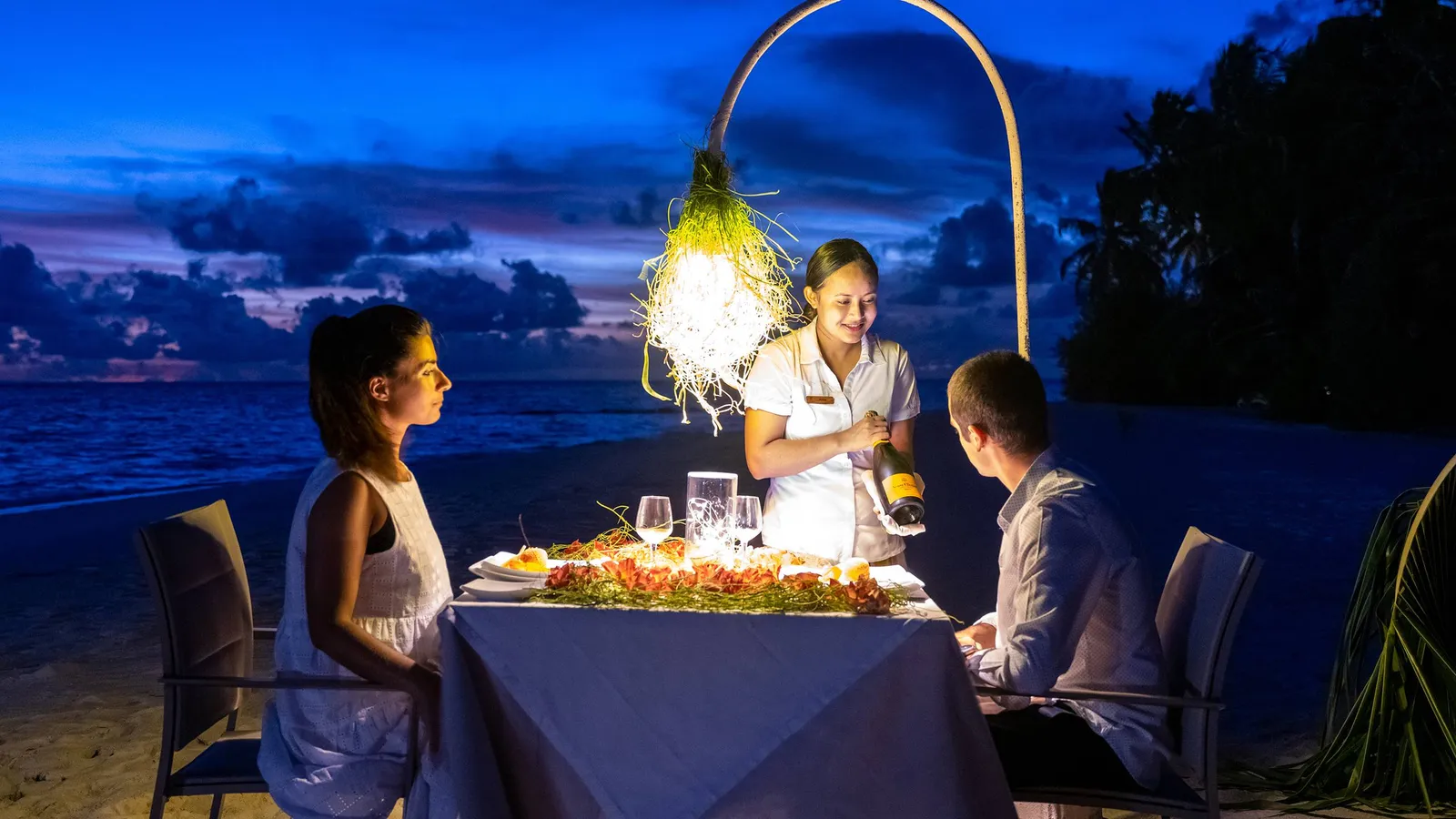 Dinner for 2 on the beach as night falls, a waitress serving lobster under a hanging lamp.