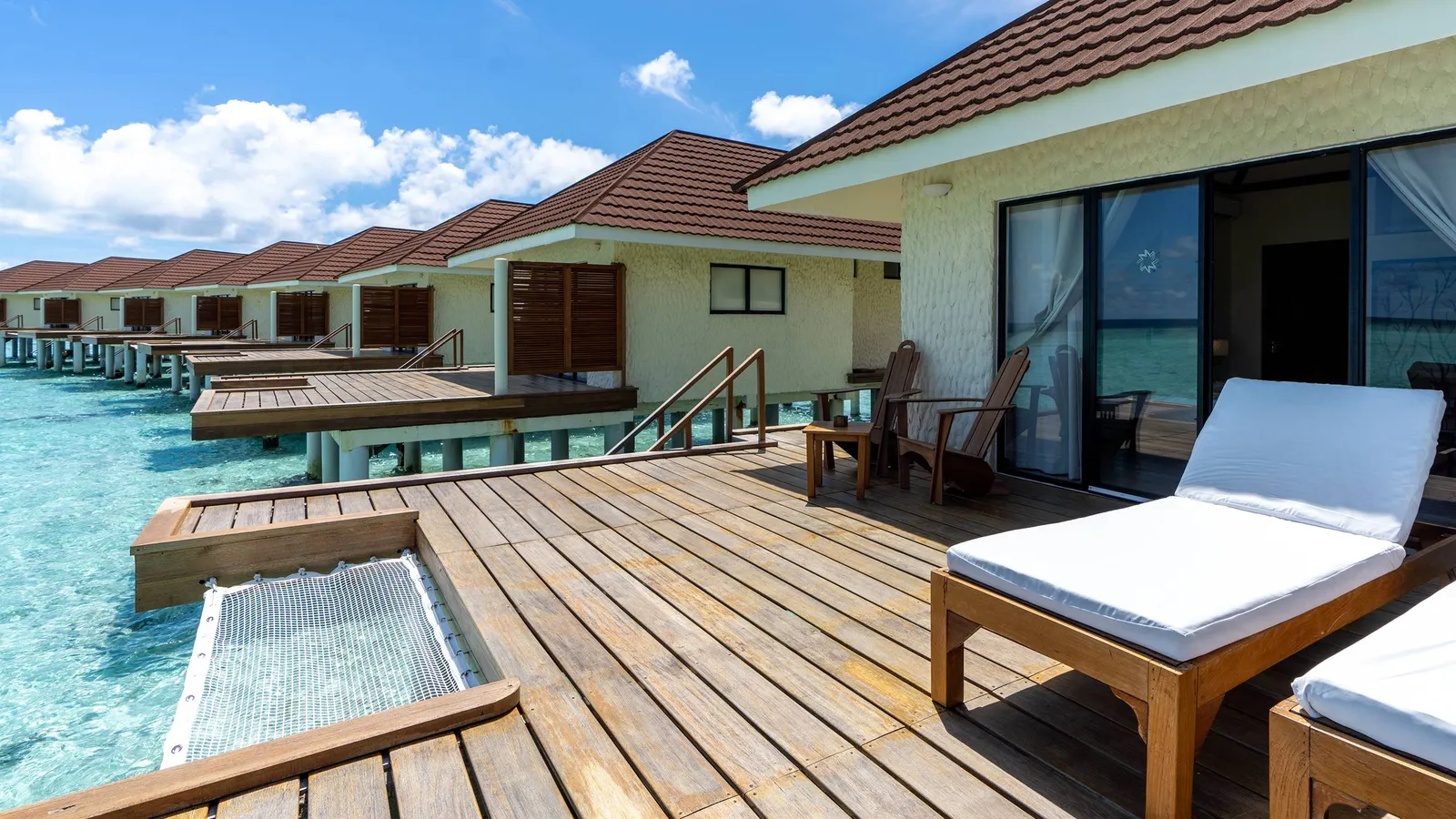 The timber deck of a water bungalow, with its lounger, the net slung over the sea and the row of bungalows beyond.