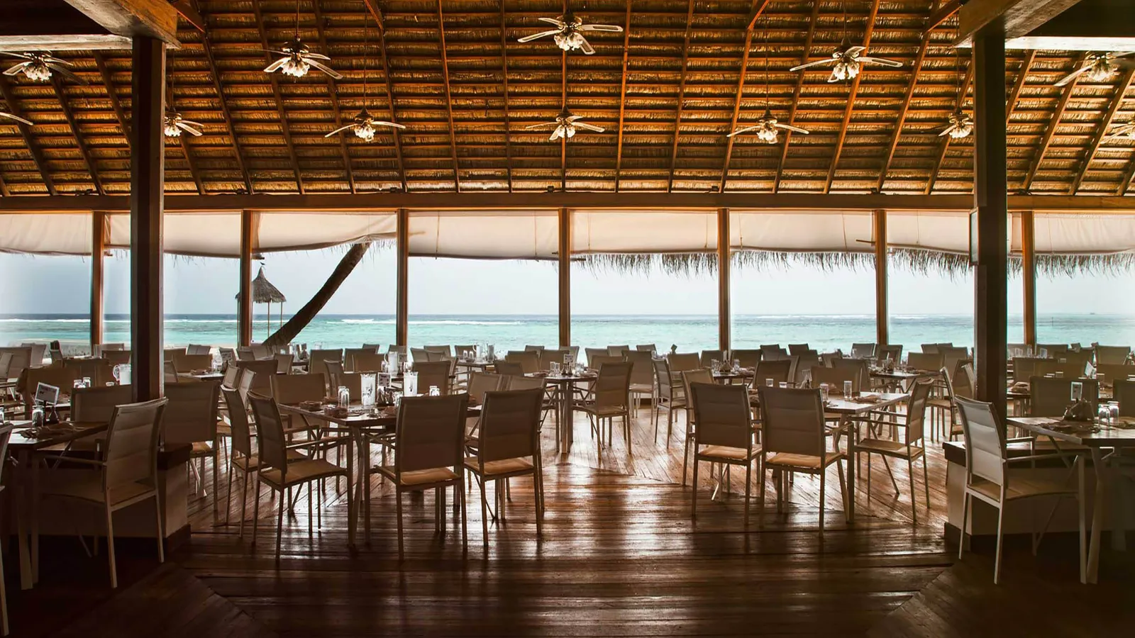 The main restaurant under its timber and thatch roof, the tables laid and the lagoon beyond the open sides.
